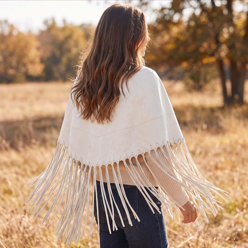 May include: A cream-colored poncho featuring a V-shaped yoke with decorative whipstitching along the seam. The garment has long, thin fringe detailing hanging from the bottom edge, creating a layered appearance over a beige sweater and dark jeans.