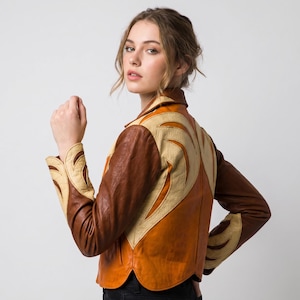 May include: A multi-toned leather jacket featuring a combination of dark brown, medium brown, and cream-colored panels. The jacket displays intricate, flame-like or wing-shaped leather appliques in cream on the sleeves and back, with a collar.