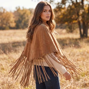 May include: A tan suede poncho featuring a wide, V-shaped hemline adorned with long, dangling fringe trim. The garment is constructed with visible whipstitch detailing along the lower edge where the fringe is attached, creating a textured appearance.