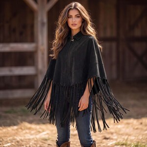 May include: A black suede poncho featuring a collared neckline, a front zipper closure, and long, thin fringe detailing along the hemline. The garment is worn over blue denim jeans, creating a layered look with a textured, matte finish.