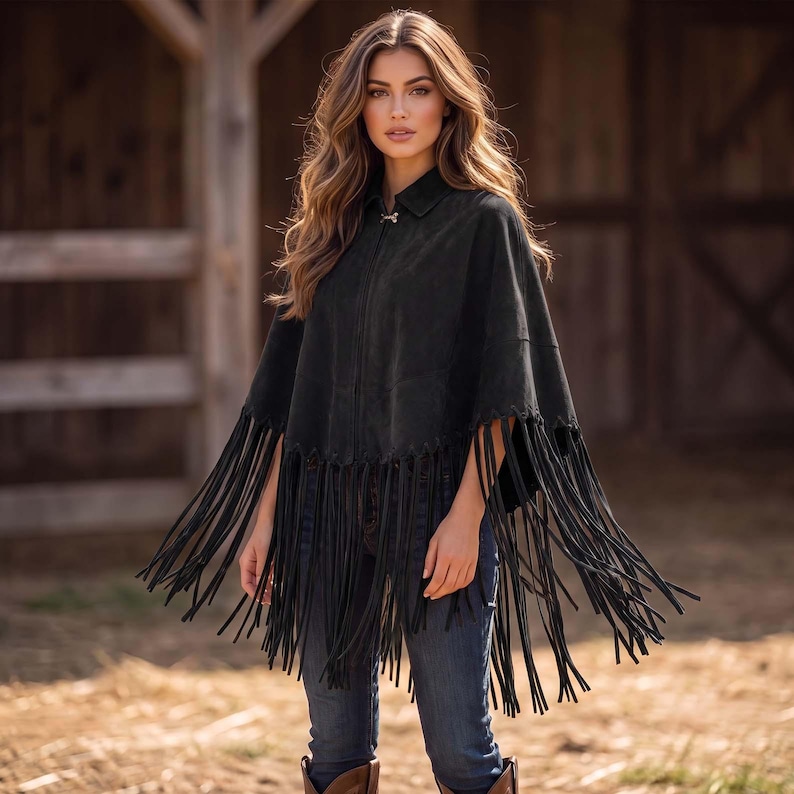 May include: A black suede poncho featuring a collared neckline, a front zipper closure, and long, thin fringe detailing along the hemline. The garment is worn over blue denim jeans, creating a layered look with a textured, matte finish.