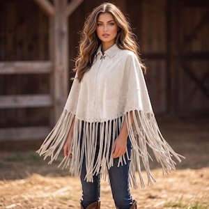 May include: A light beige suede poncho featuring a collared neckline, a full-length front zipper, and long fringe detailing along the hemline. The garment is worn over dark blue denim jeans, creating a layered look with a textured finish.
