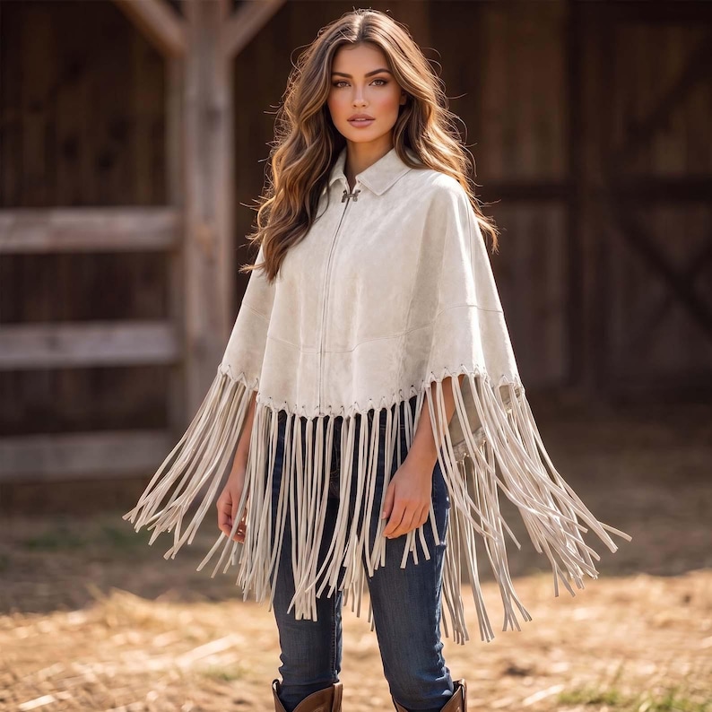 May include: A light beige suede poncho featuring a collared neckline, a full-length front zipper, and long fringe detailing along the hemline. The garment is worn over dark blue denim jeans, creating a layered look with a textured finish.