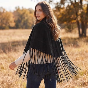 May include: A black suede cape featuring a V-shaped hemline adorned with long, thin fringe detailing along the entire bottom edge. The garment is worn over a cream-colored long-sleeved ribbed sweater and dark blue denim jeans.