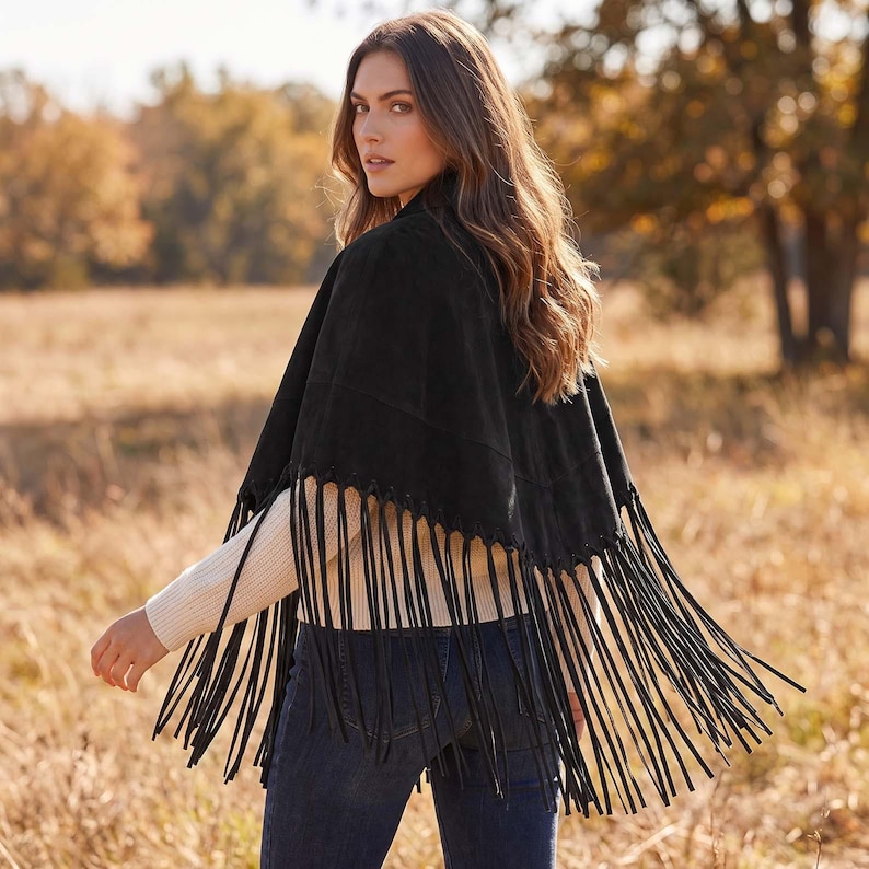 May include: A black suede cape featuring a V-shaped hemline adorned with long, thin fringe detailing along the entire bottom edge. The garment is worn over a cream-colored long-sleeved ribbed sweater and dark blue denim jeans.