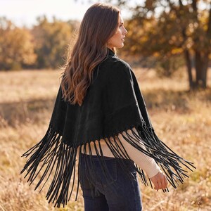 May include: A black suede poncho featuring a V-shaped hemline adorned with long, thin black fringe trim. The garment is worn over a cream-colored long-sleeved ribbed sweater and dark blue denim jeans, showcasing a layered look from the back.
