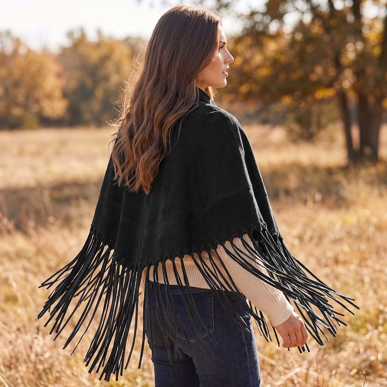 May include: A black suede poncho featuring a V-shaped hemline adorned with long, thin black fringe trim. The garment is worn over a cream-colored long-sleeved ribbed sweater and dark blue denim jeans, showcasing a layered look from the back.