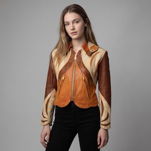 May include: A multi-toned leather jacket featuring a color-blocked design in shades of cream, tan, and dark brown. The jacket has a central gold-tone zipper, two zippered pockets on the front, a pointed collar, and decorative curved paneling.