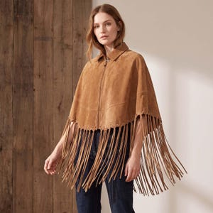 May include: A tan suede cape featuring a front zipper closure, a pointed collar, and a wide hemline adorned with long, dangling fringe trim. The garment is worn over dark blue denim jeans, showcasing a textured finish and a structured silhouette.