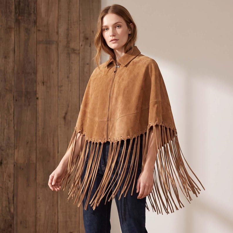 May include: A tan suede cape featuring a front zipper closure, a pointed collar, and a wide hemline adorned with long, dangling fringe trim. The garment is worn over dark blue denim jeans, showcasing a textured finish and a structured silhouette.