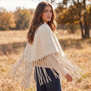 May include: A cream-colored suede poncho featuring a V-shaped hemline with long, dangling fringe trim. The garment includes a woven knit texture on the sleeves and is worn over dark blue denim jeans, showcasing a layered and textured appearance.