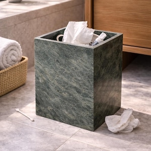 May include: A square, dark green marble waste bin in a bathroom. The bin contains tissues and a small bottle. A rolled white towel is in a wicker basket to the left. A cotton bud lies on the floor.