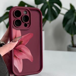 May include: A burgundy phone case with a pink lily flower design. The case has a smooth finish and a raised edge around the camera lenses. The floral design is centered on the back of the case.