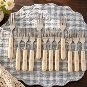 May include: A set of twelve stainless steel forks with cream-colored handles featuring a delicate pink floral pattern. The forks are arranged in rows on a blue and white checkered quilted fabric mat, showcasing a consistent design across all pieces.