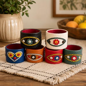 May include: A set of six cylindrical napkin rings made of textured leather in black, white, blue, orange, red, and green. Each ring features a colorful stitched eye or heart motif decorated with small, round, multi-colored buttons.