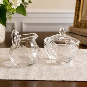May include: A clear glass creamer and a matching sugar bowl with a lid, both featuring an etched leaf pattern. The creamer has a curved handle and a pouring spout, while the round sugar bowl is topped with a small, pointed glass finial.