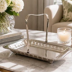 May include: A rectangular silver-toned serving tray with a polished reflective surface, a beaded rim, and a single arched handle attached to the centre of the long sides. The tray is supported by four small feet and features a smooth, metallic finish.