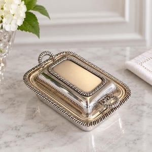 Peut inclure: Un beurrier rectangulaire de couleur argentée avec un couvercle assorti, doté d'une finition métallique polie et d'une bordure décorative en forme de corde. Le couvercle comprend un panneau central rectangulaire et deux petites anses incurvées.