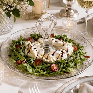 Könnte beinhalten: Eine Servierplatte aus klarem Glas mit einem mittigen Griff und einem dekorativen, eingravierten Blattmuster am Rand. Die Platte ist mit einem Bett aus Rucola, halbierten Kirschtomaten und weißem Käse belegt, der mit Balsamico-Creme beträufelt ist.