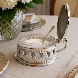 Puede incluir: Un azucarero de metal plateado con un patrón de filigrana ornamentado y una tapa abatible. El soporte contiene un cuenco de cristal transparente facetado lleno de azúcar blanca e incluye una pequeña cuchara decorativa de metal plateado.
