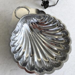 May include: A shiny, silver-colored serving dish in the shape of a seashell. The dish has a scalloped edge and a handle. A decorative metal piece with floral accents is in the background. The dish is likely made of metal.