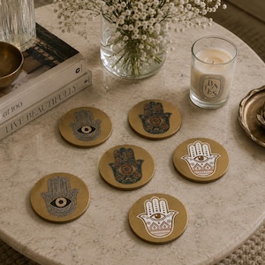 May include: A set of six circular gold-toned coasters, each featuring a distinct Hamsa hand design. The designs include intricate black line art, detailed multi-coloured patterns, and bold white graphic prints, all centred with an eye motif.