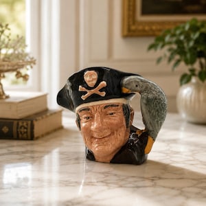 May include: A ceramic character jug shaped like a pirate wearing a black tricorn hat with a white skull and crossbones emblem. The pirate has a tan complexion and a subtle smile. A grey parrot is perched on the side, forming the handle of the jug.