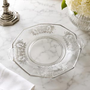 May include: A clear glass serving tray with an octagonal shape and two integrated side handles. The tray features a decorative silver-coloured floral pattern printed along the rim, showcasing intricate botanical details and scrolling foliage.