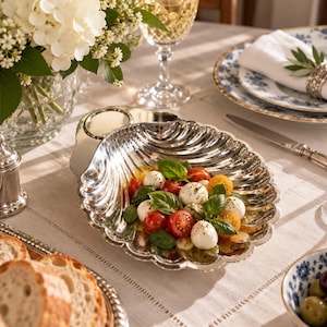 May include: A polished silver serving dish shaped like a large scallop shell, containing a salad of red and yellow cherry tomatoes, white mozzarella balls, and fresh green basil leaves, arranged on a table with other dining tableware.