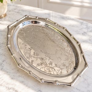 May include: An oval silver-coloured serving tray featuring a raised bamboo-style rim and an intricate engraved pattern on the base depicting delicate flowering branches and small butterflies. The polished metallic surface reflects light across the design.