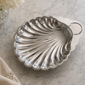 Puede incluir: Un plato de metal plateado pulido con forma de concha de vieira. El plato presenta profundas ranuras estriadas y radiantes que se extienden desde una zona plana circular y lisa en el borde superior hacia el borde inferior curvo.