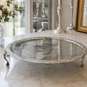 May include: A round silver-toned metal serving tray with a reflective surface and an ornate, embossed floral pattern along the raised rim. The tray is supported by three curved, decorative legs that elevate it above the surface.