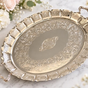 May include: An oval silver-coloured serving tray featuring ornate embossed floral patterns along the border and a central decorative engraving. The tray includes two intricate, curved handles on opposite ends and a smooth, reflective centre surface.
