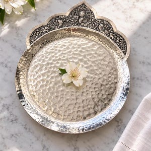 Puede incluir: Una bandeja redonda de metal plateado con una textura martillada en el centro. El borde superior presenta un adorno elevado con grabados intrincados de flores y pájaros. Una sola flor blanca descansa en el centro de la bandeja.