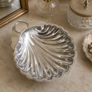 May include: A polished silver-toned metal dish shaped like a scallop shell. The dish features a smooth, reflective surface with deep, radiating fluted grooves and a small circular handle attached to the top edge, creating a decorative tray.