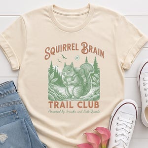 May include: Cream-colored t-shirt featuring a green illustration of a squirrel with a backpack, set against a forest backdrop. The text "Squirrel Brain Trail Club" is arched above the squirrel, with "Powered by Snacks and Side Quests" below.