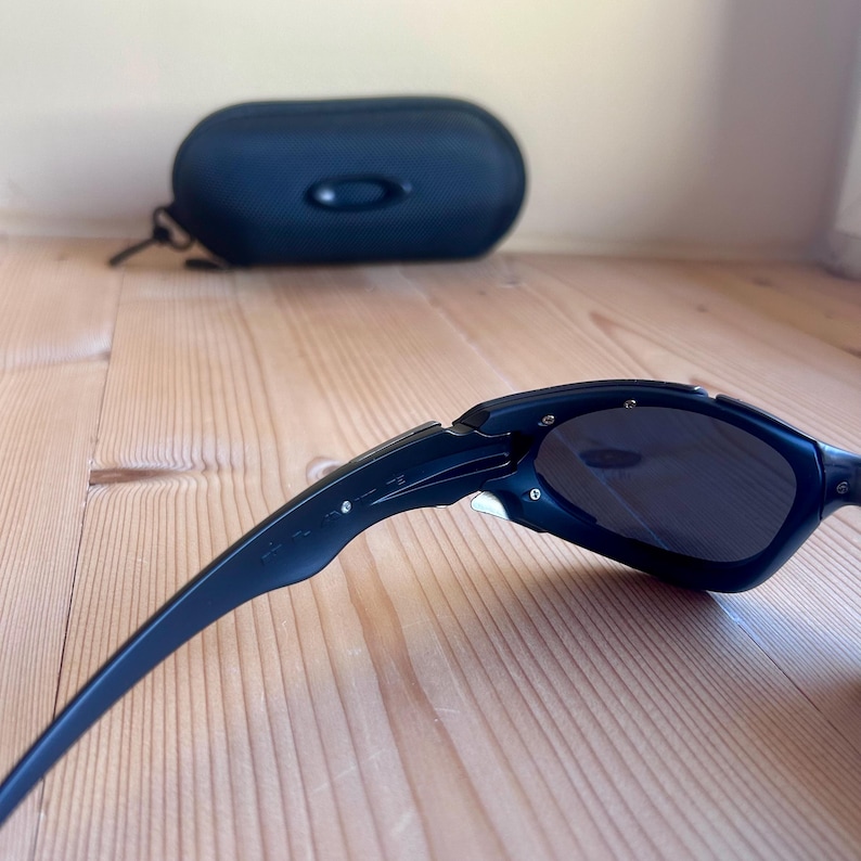 May include: Black sunglasses with dark lenses, angled on a wooden surface. A black, oval-shaped case is in the background. The sunglasses have a sleek, modern design, with a curved frame and a small logo on the arm.
