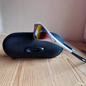 May include: Silver-framed sunglasses with mirrored lenses, resting on a black, oval-shaped case. The lenses reflect yellow, red, and blue hues. The case has a textured surface and a black oval logo.