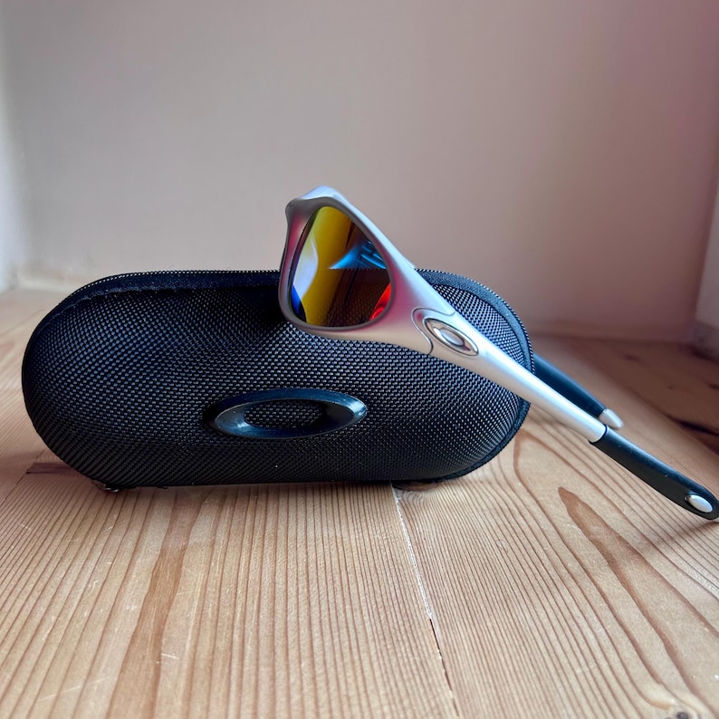 May include: Silver-framed sunglasses with mirrored lenses, resting on a black, oval-shaped case. The lenses reflect yellow, red, and blue hues. The case has a textured surface and a black oval logo.