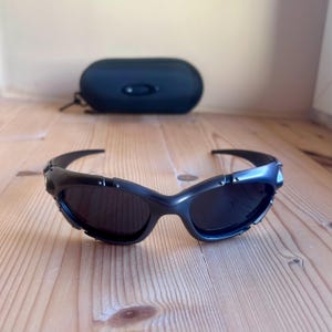 May include: Black sunglasses with a curved frame and dark lenses. The sunglasses are on a wooden surface, with a black case in the background. The sunglasses have a sporty, modern design.