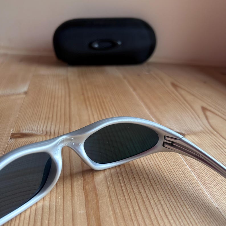 May include: Silver-framed sunglasses with dark lenses rest on a wooden surface. A black sunglasses case is in the background. The sunglasses have a sleek, modern design.
