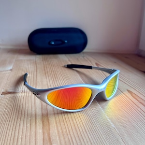 May include: Silver-framed sunglasses with vibrant orange and yellow lenses rest on a wooden surface. A dark blue case is in the background. The sunglasses have a sleek, modern design.