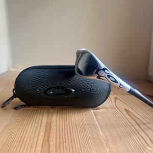 May include: White and black sunglasses with a decorative flame design, resting on a black, oval-shaped case. The sunglasses have dark lenses and a sleek, modern design. The case has a zipper and a small, oval logo.