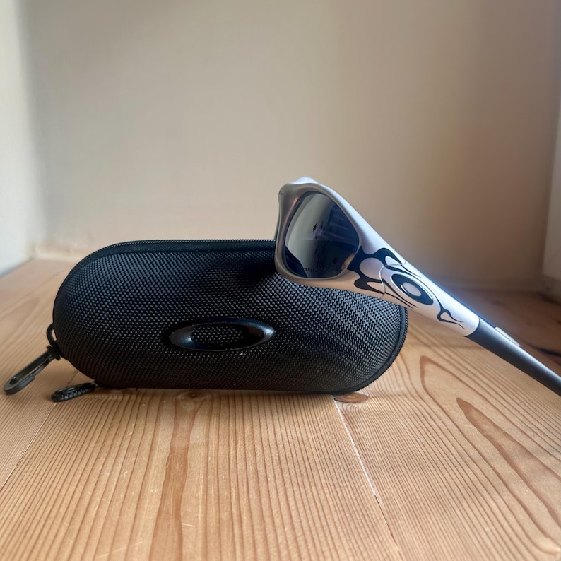 May include: White and black sunglasses with a decorative flame design, resting on a black, oval-shaped case. The sunglasses have dark lenses and a sleek, modern design. The case has a zipper and a small, oval logo.