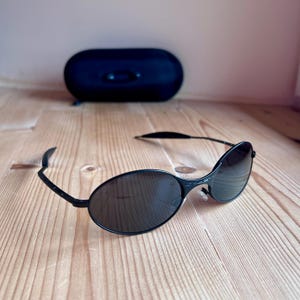May include: Black oval sunglasses with a matching black case. The sunglasses have a sleek, modern design with dark lenses and a thin frame. The case is oval-shaped and sits in the background on a wooden surface.