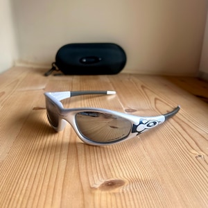 May include: Silver sunglasses with mirrored lenses and black and white detailing on the arms. A black case is visible in the background. The sunglasses are displayed on a wooden surface.