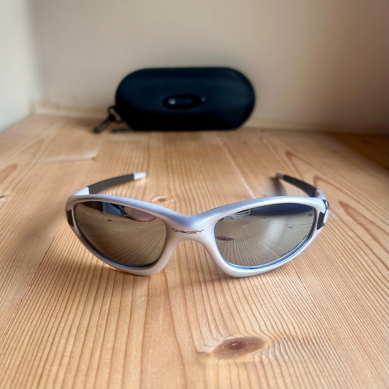 May include: Silver and black Oakley sunglasses with mirrored lenses. The sunglasses are on a wooden surface, with a black case in the background. The Oakley logo is visible on the frame.