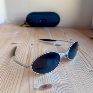 Puede incluir: Gafas de sol con montura plateada y lentes oscuras sobre una superficie de madera. Las gafas tienen un diseño elegante con patillas curvas y una funda negra en el fondo. Las lentes son de forma ovalada.
