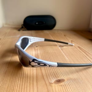 May include: A pair of white and black sunglasses with a decorative design on the frame. The sunglasses have dark lenses and black arms. A black sunglasses case is in the background.