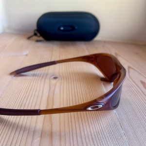 Op de afbeelding: Bruine zonnebril met bijpassende etui. De zonnebril heeft een gebogen montuur met donkerbruine pootjes en glazen. De etui is zwart en ovaal van vorm.